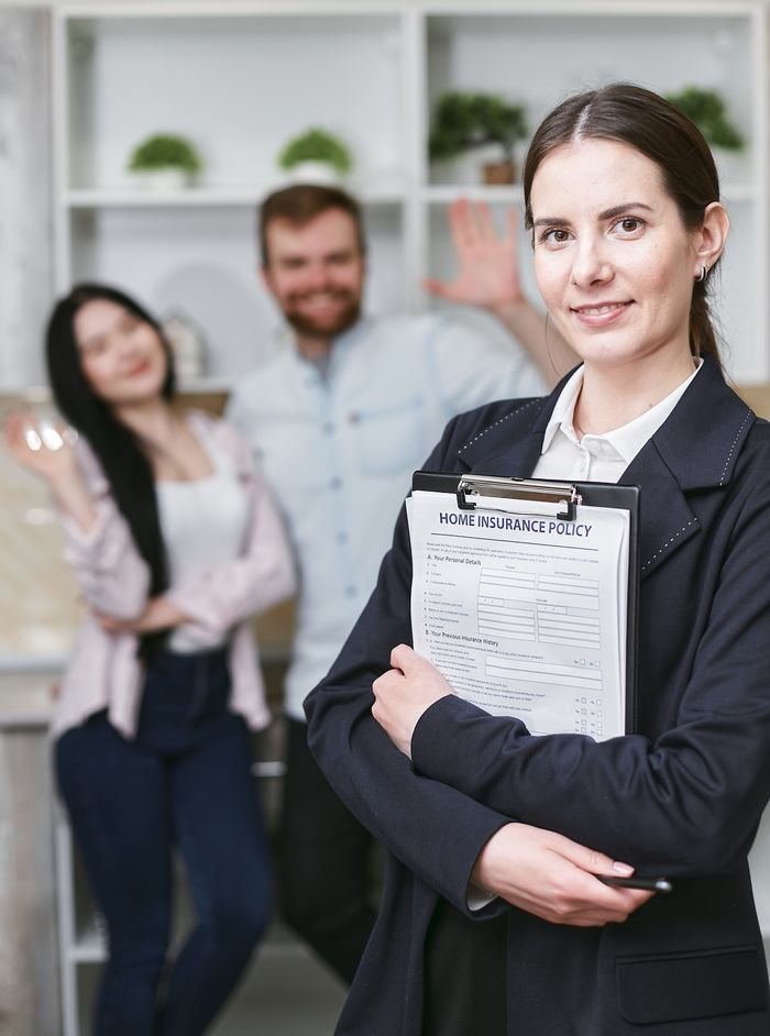 Home A smiling businesswoman holding a home insurance policy with clients in the background.